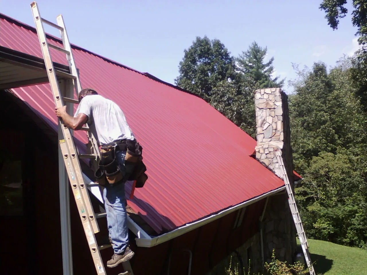 Roofer on a ladder beside a red metal roof and stone chimney — Soffit & Fascia Repair by Roofing Brothers in Virgin